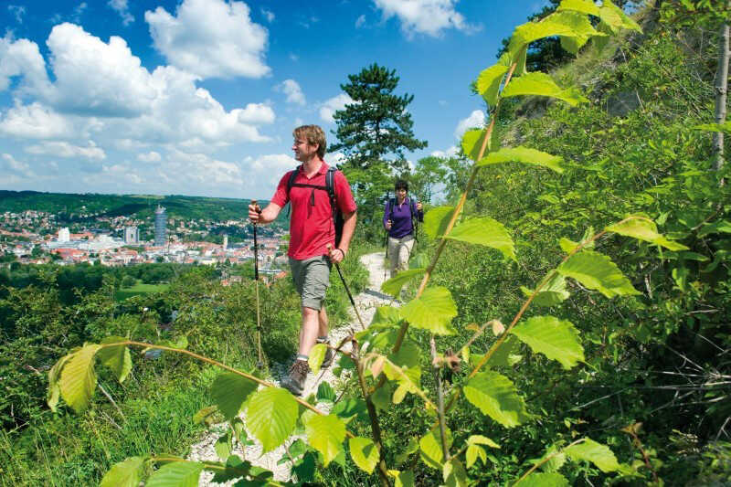 Zwei Wanderer genießen auf einem sonnigen Höhenweg den weiten Blick über Jena und die grüne Saalelandschaft.