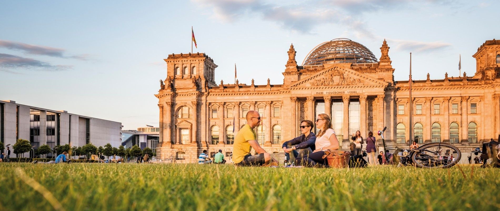 Eine Gruppe von Menschen sitzt auf einer Wiese vor dem Reichstagsgebäude in Berlin bei sonnigem Wetter.