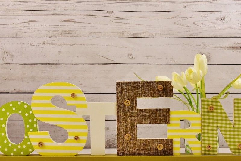 Dekorativer Schriftzug 'OSTERN' in verschiedenen Materialien und Mustern, neben gelben Tulpen vor einer Holzwand.