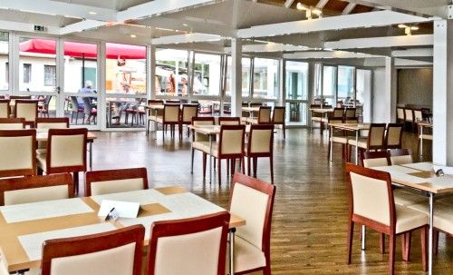 Ein geräumiges, modern eingerichtetes Restaurant mit Holzboden, leeren Tischen und Stühlen. Große Fenster lassen viel Tageslicht herein.