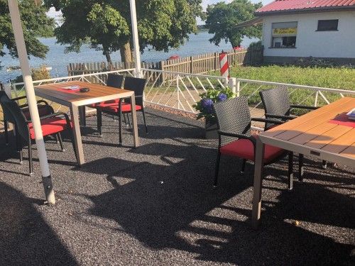 Sitzbereich eines Cafés mit Holztischen und geflochtenen Stühlen, Blick auf einen See im Hintergrund.