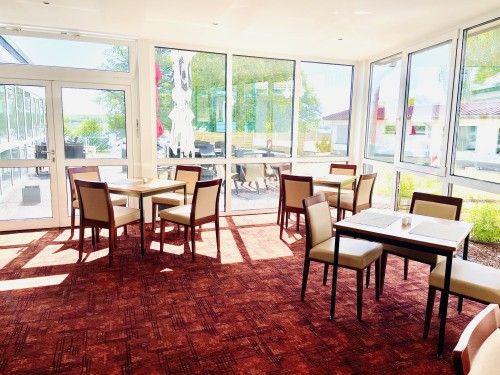 Heller, leerer Essbereich mit mehreren Tischen und Stühlen, großen Fenstern und Blick nach draußen.