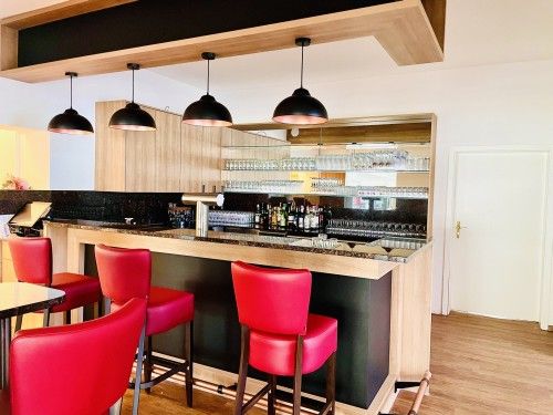 Moderne Bar mit Holzverkleidung, roten Barhockern und Hängelampen. Gläser und Flaschen sind im Hintergrund zu sehen.