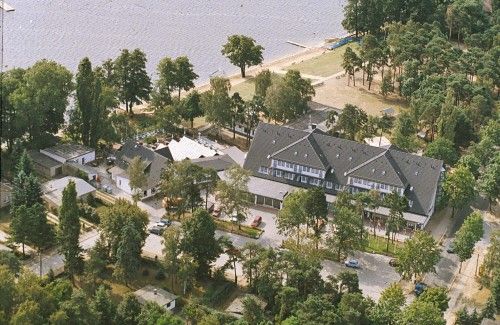 Luftaufnahme eines großen Gebäudekomplexes umgeben von Bäumen in der Nähe eines Sees mit Strand.