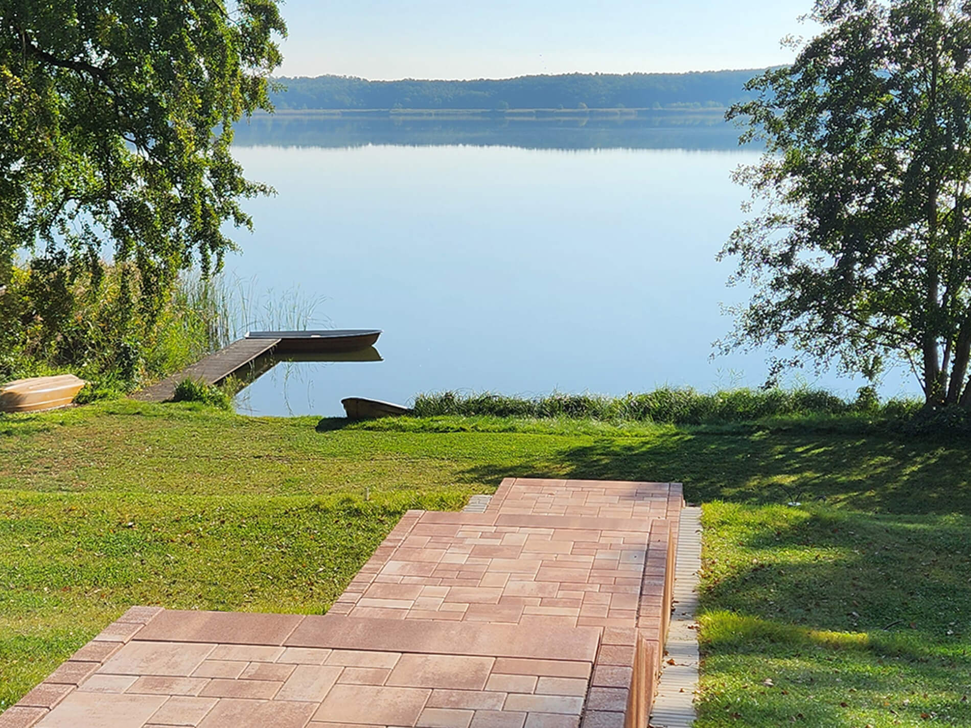 Ein Holzsteg führt zum See, umrahmt von Bäumen, während die Sonne den Himmel in lebhaften Farben bei Sonnenaufgang erleuchtet.