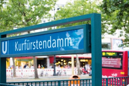 Eingang zum U-Bahnhof Kurfürstendamm mit einem blauen Schild, Bäume und ein vorbeifahrender roter Bus im Hintergrund