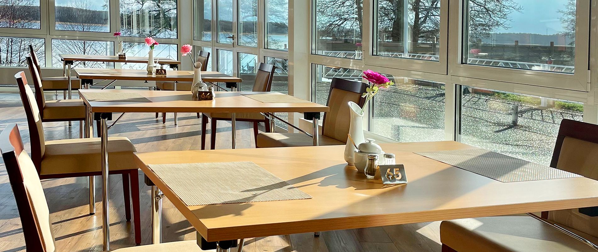 Helles Restaurant mit großen Fenstern, mehreren Tischen mit Blumen und Blick auf einen sonnigen Außenbereich.