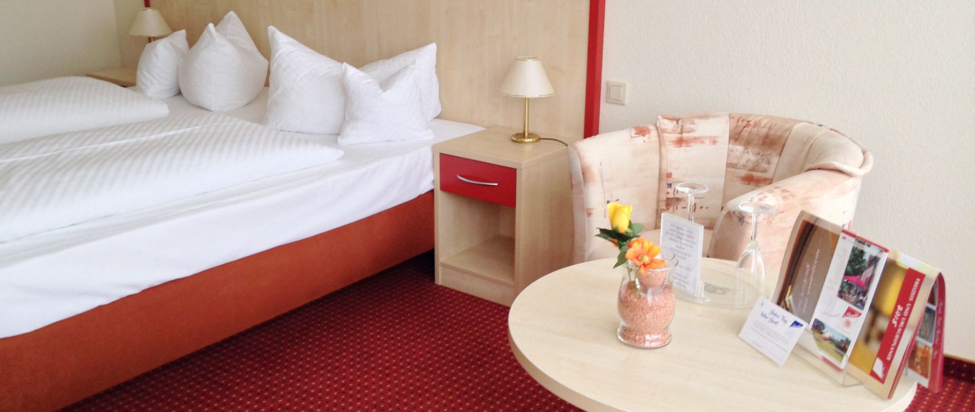 Hotelzimmer mit Doppelbett, Nachttisch, runder Tisch, Sessel und Broschüren